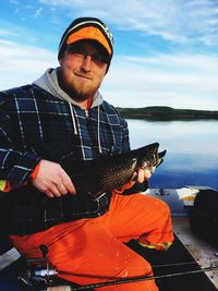 Portrait of young man holding fish while sitting on boat on lake against sky