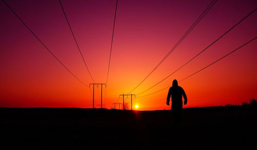 Silhouette man standing by electricity pylon against sky during sunset