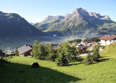 Trees and grass on field against houses and mountains during sunny day