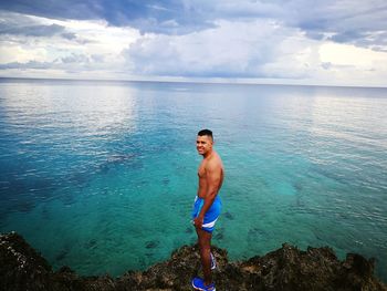 Portrait of shirtless man on rock by sea against sky