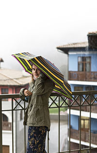  young woman in khaki jacket with umbrella in rainbow pattern in drops standing on terrace in rain 