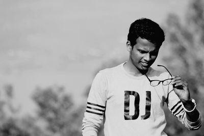 Young man holding eyeglasses standing against sky