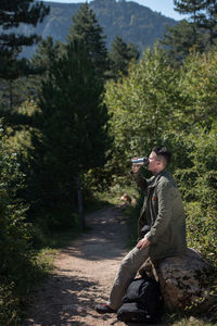 Side view of man photographing while sitting on tree