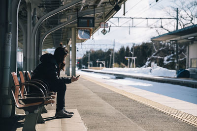 Side view of woman using smart phone sitting on bench at railroad station