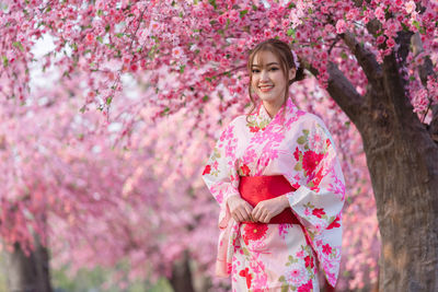 Portrait of smiling woman standing by pink cherry blossom tree