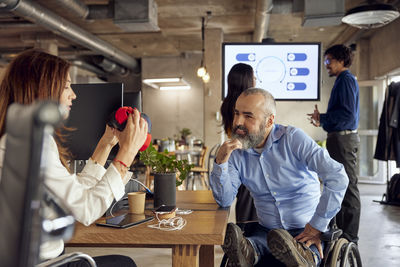 Smiling businessman with disability looking at colleague discussing over vr simulator in creative office