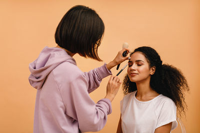 Beautician applying make-up on woman against beige background