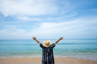 Rear view of carefree woman with arms raised standing at beach against sky