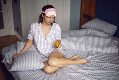 Woman in pajamas and with a sleep mask on the bed in the bedroom and drinking orange juice 