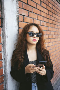 Young woman using phone while standing against brick wall