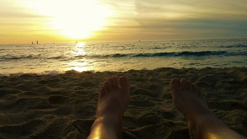 Low section of woman relaxing on beach against sky during sunset