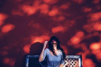 Woman holding flower in mouth while sitting on bench against wall