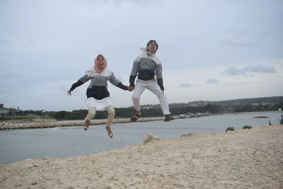 Full length of friends holding hands while jumping at beach against sky