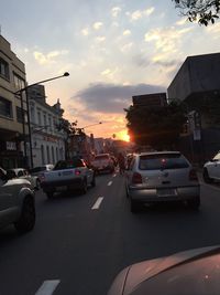 Cars on road by buildings against sky during sunset