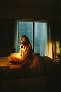 Side view of young female traveler in white shirt and sunglasses with towel wrapped around head sitting on comfortable bed and talking on telephone in modern hotel room