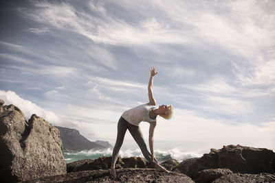 Woman practicing trikonasana on rock formation against sky