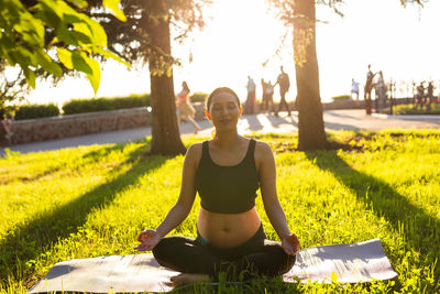 Full length of woman meditating while sitting on grass against trees