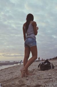 Full length of woman standing at beach by shoulder bag against sky during sunset