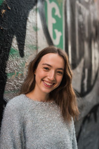 Positive young female leaning on wall with colorful creative graffiti while standing in urban area and looking at camera