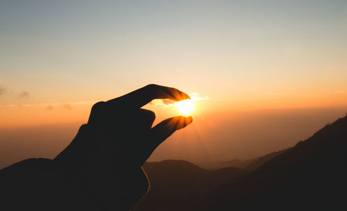 Optical illusion of silhouette person holding sun over mountains during sunset