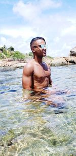 Shirtless young man wearing sunglasses swimming in sea against cloudy sky