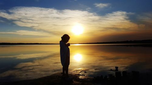 Silhouette of a girl standing on shore against sky during sunset