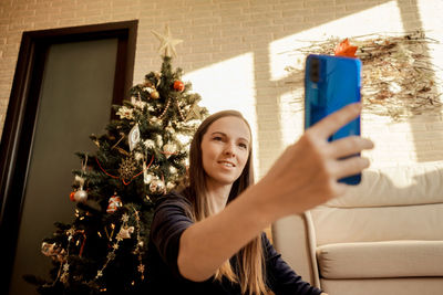 Smiling woman doing selfie while sitting against christmas tree