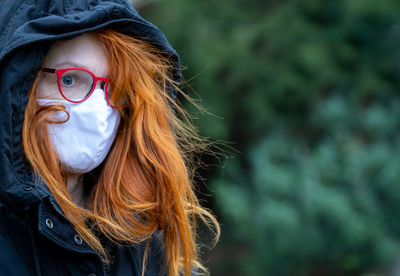 A hooded woman with red hair stands lonely and sad in a park. she wears a breathing mask.