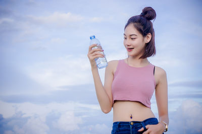 Portrait of smiling young woman with water bottle standing against cloudy sky