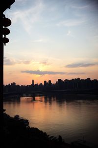 Silhouette buildings by river against sky during sunset