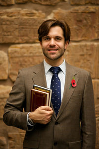 Portrait of smiling man holding books while standing against wall