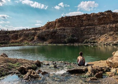 Man sitting on rock by lake against sky