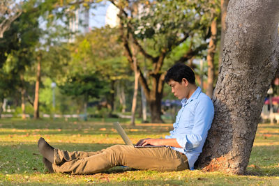 Side view of man wearing mask using laptop while sitting by tree trunk at park