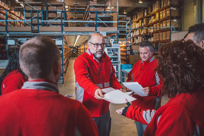 Bearded senior male manager giving instructions on documents to group of employees in red uniform i modern warehouse