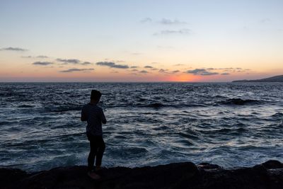 Man standing on rocks at beach against sky during sunset