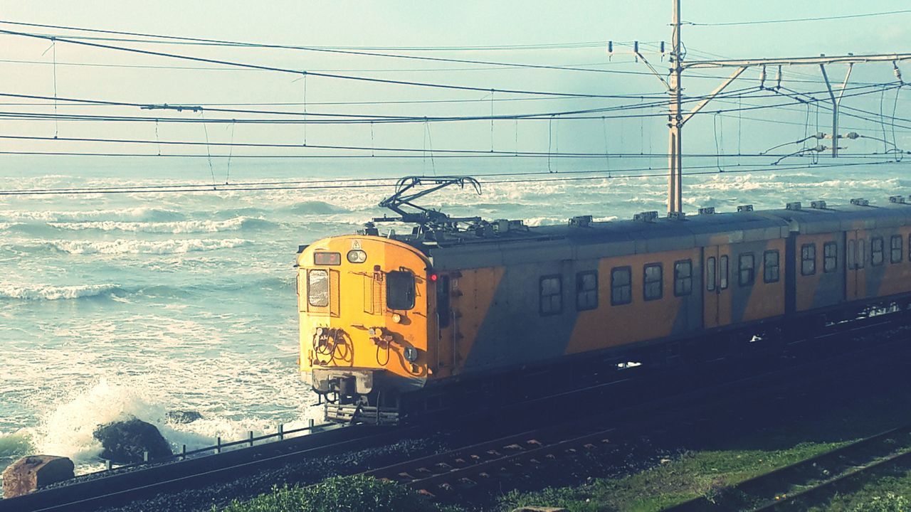 transportation, railroad track, rail transportation, public transportation, built structure, architecture, railroad station platform, mode of transport, train - vehicle, power line, building exterior, railroad station, sky, connection, yellow, land vehicle, road, electricity pylon, outdoors, street