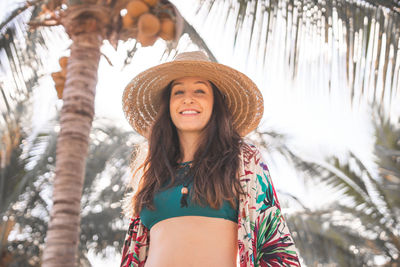 Portrait of young woman wearing hat standing against palm trees