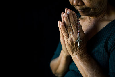 Midsection of senior woman holding rosary praying against black background