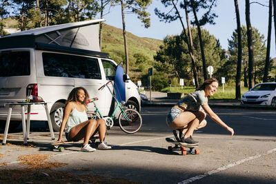 Beautiful skater practicing riding skate board on street with background of woman sitting on skate near minivan in camping