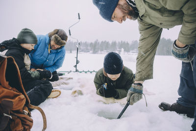 Boy looking at father ice fishing while friends sitting on snow during vacation