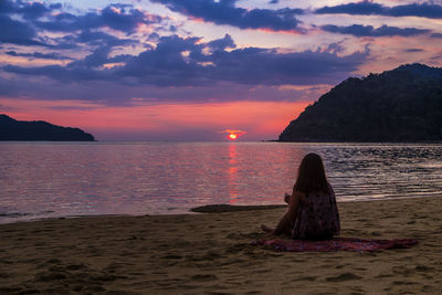 Rear view of woman sitting at beach against sky during sunset