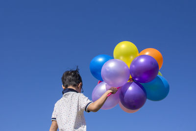 Low angle rear view of boy holding multi colored balloons against clear blue sky