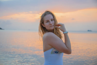 Portrait of young woman standing at beach against sky during sunset