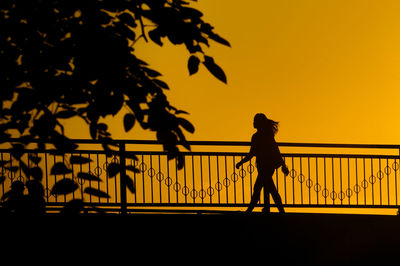 Silhouette woman walking by railing against orange sky