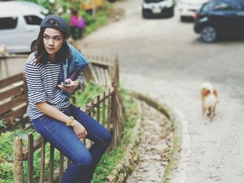 Young woman using phone while sitting on fence at roadside