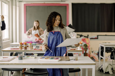 Teenage female student examining painting standing near desk during art class at high school