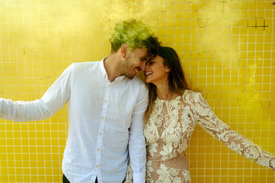 Young romantic newlywed couple in stylish wedding outfits having fun with yellow smoke bombs while standing together near modern building in tropical city