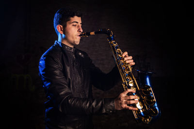 Side view of young man playing saxophone against black background