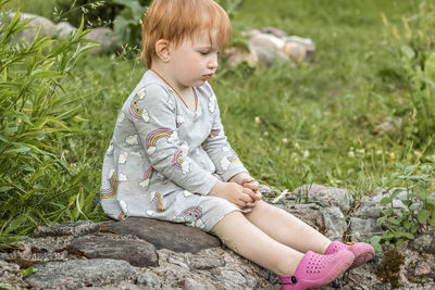 Little girl with red hair sits with a camomile in her hand in the garden among the greenery.