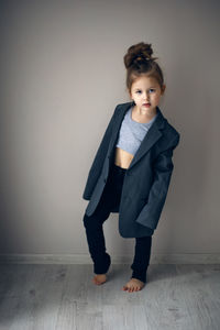 Funny portrait of fashionable girl child in large gray jacket stands against gray wall next  window 
